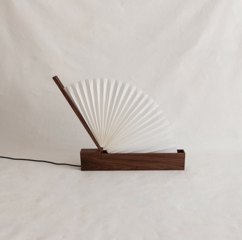 A lamp with a pleated, fan-shaped white shade and a wooden base, connected to a black power cord, set against a plain background