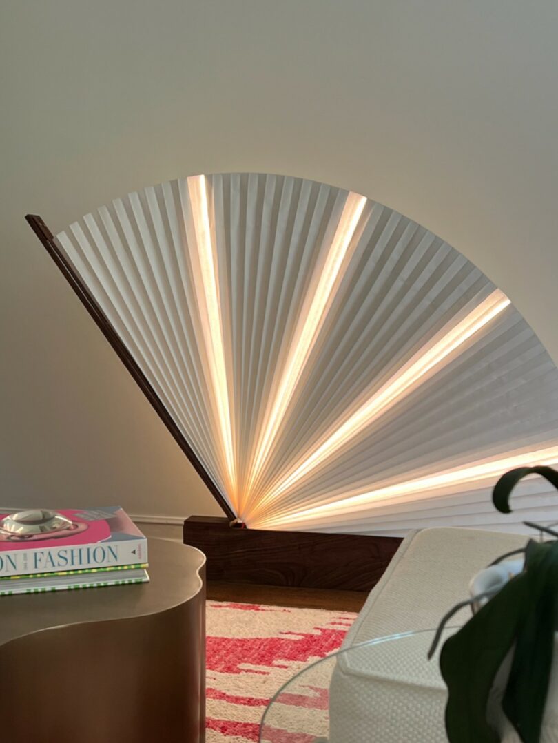 A modern fan-shaped lamp with pleated white material and warm light sits on the floor beside a round gold table with books and a patterned rug