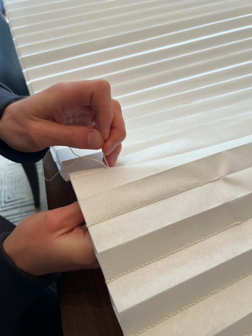 Hands sewing the edge of a white pleated fabric or paper material with a needle and thread on a wooden surface