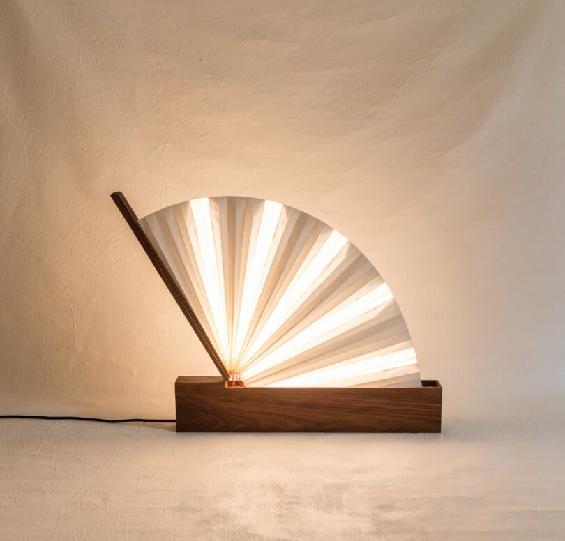 A lamp with a wooden rectangular base and a fan-shaped paper shade emits warm light, positioned against a plain, textured wall