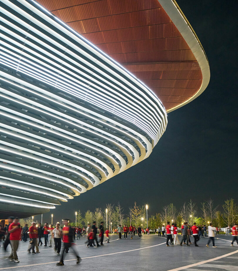 People walk outside a large, modern stadium at night; the stadium features curved, illuminated horizontal lines and a wooden overhang.