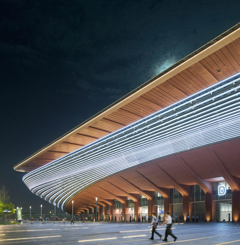 A large, modern building with curved, illuminated roof lines is shown at night. People walk near the entrance beneath a sign marked “D.” The sky is dark with clouds.