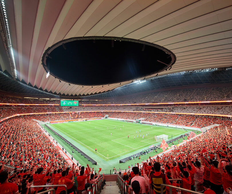 A packed stadium with fans in red watches a soccer match on a well-lit field, with players actively engaged in play under a large, open circular roof.