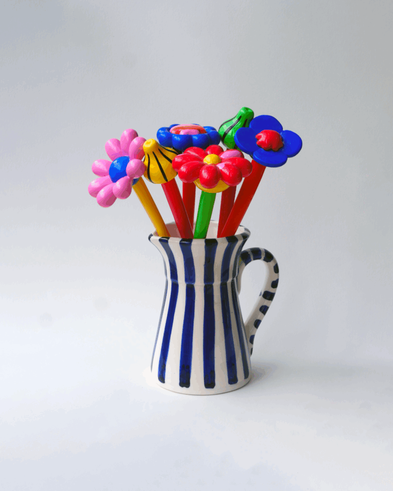 A white ceramic pitcher with blue vertical stripes holds several colorful pens topped with flower designs, against a plain light background