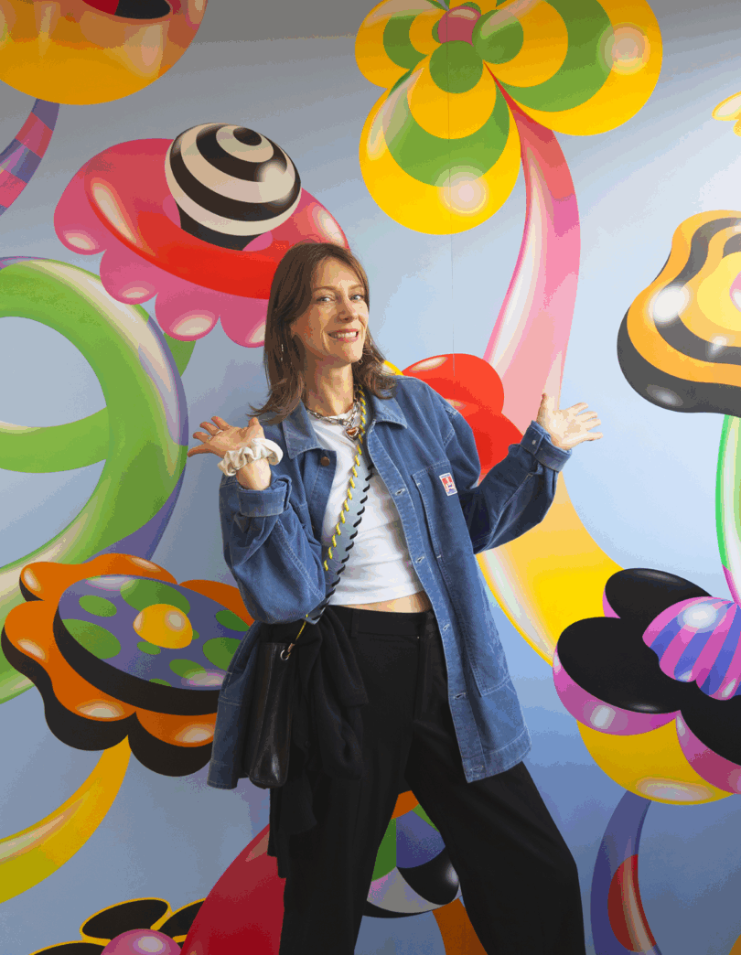 A woman in a denim jacket stands smiling with arms raised in front of a colorful, abstract mural featuring bright, whimsical shapes and patterns