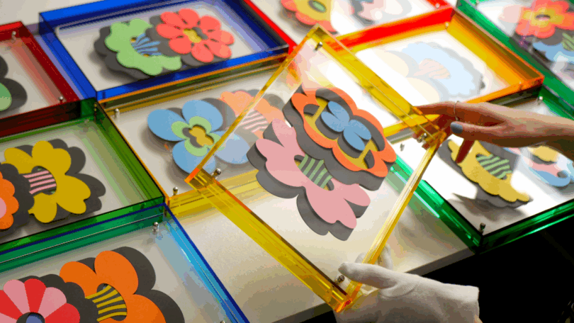 A person wearing gloves arranges colorful layered flower artworks enclosed in transparent plastic frames on a table